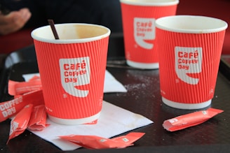 Three red paper cups with the 'Café Coffee Day' logo are placed on a tray. There are also several empty sugar sachets scattered around on white napkins.