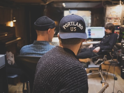 Three people are in a recording studio. Two individuals sit facing away, looking at a third person in the background who is seated in front of a computer screen with audio editing software open. The person in focus wears a dark sweater and a navy cap with 'PORTLAND US' written on it. The atmosphere suggests collaboration and music production.