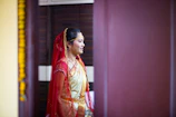 A person is dressed in traditional attire, featuring a red and gold saree with intricate details. The individual has a red veil draped over their head and is adorned with jewelry, including a necklace and earrings. The setting appears to be indoors, with a dark brown background and a hint of yellow decoration visible on the left side.