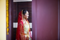 A person is dressed in traditional attire, featuring a red and gold saree with intricate details. The individual has a red veil draped over their head and is adorned with jewelry, including a necklace and earrings. The setting appears to be indoors, with a dark brown background and a hint of yellow decoration visible on the left side.