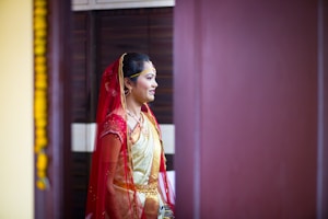 A person is dressed in traditional attire, featuring a red and gold saree with intricate details. The individual has a red veil draped over their head and is adorned with jewelry, including a necklace and earrings. The setting appears to be indoors, with a dark brown background and a hint of yellow decoration visible on the left side.