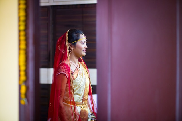 A person is dressed in traditional attire, featuring a red and gold saree with intricate details. The individual has a red veil draped over their head and is adorned with jewelry, including a necklace and earrings. The setting appears to be indoors, with a dark brown background and a hint of yellow decoration visible on the left side.