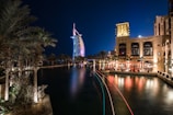 A night scene featuring an illuminated, iconic sail-shaped building with colorful lights reflecting on a tranquil waterway. Palm trees are lining the water, and a luxurious resort is visible with people dining at a waterfront terrace, casting a warm glow.