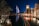 A night scene featuring an illuminated, iconic sail-shaped building with colorful lights reflecting on a tranquil waterway. Palm trees are lining the water, and a luxurious resort is visible with people dining at a waterfront terrace, casting a warm glow.