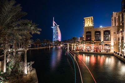 A night scene featuring an illuminated, iconic sail-shaped building with colorful lights reflecting on a tranquil waterway. Palm trees are lining the water, and a luxurious resort is visible with people dining at a waterfront terrace, casting a warm glow.
