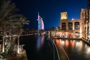 A night scene featuring an illuminated, iconic sail-shaped building with colorful lights reflecting on a tranquil waterway. Palm trees are lining the water, and a luxurious resort is visible with people dining at a waterfront terrace, casting a warm glow.