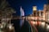 A night scene featuring an illuminated, iconic sail-shaped building with colorful lights reflecting on a tranquil waterway. Palm trees are lining the water, and a luxurious resort is visible with people dining at a waterfront terrace, casting a warm glow.