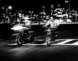Two motorcyclists are riding on a road with a cityscape in the background at night. The image is in black and white, with an emphasis on the motion blur of the vehicles, creating a dynamic and energetic feel. Bright lights and reflections add to the urban atmosphere.