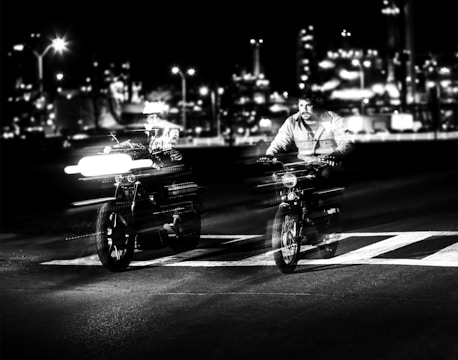 Two motorcyclists are riding on a road with a cityscape in the background at night. The image is in black and white, with an emphasis on the motion blur of the vehicles, creating a dynamic and energetic feel. Bright lights and reflections add to the urban atmosphere.