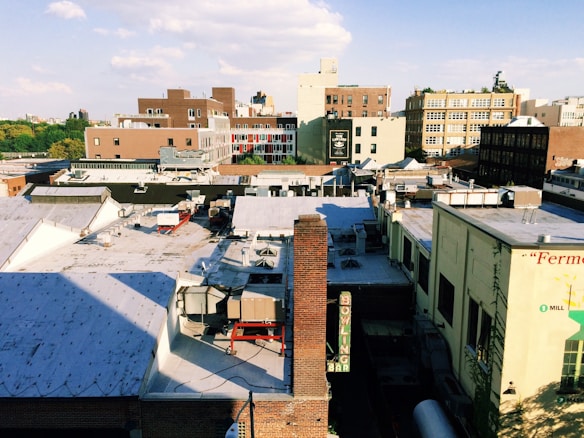 An urban rooftop landscape featuring a mix of low-rise buildings and rooftops with HVAC units and chimneys. In the background, taller buildings with a blend of neutral and brick tones are visible. The sky is partly cloudy with ample sunlight casting shadows over the scene.
