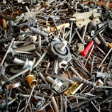 A chaotic assortment of various metal and plastic hardware items, including screws, bolts, anchors, a hinge, and other fasteners. The objects overlap each other, creating a dense and intricate texture.