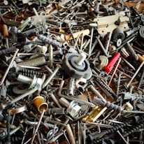 A chaotic assortment of various metal and plastic hardware items, including screws, bolts, anchors, a hinge, and other fasteners. The objects overlap each other, creating a dense and intricate texture.