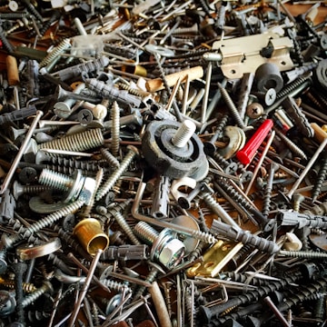 A chaotic assortment of various metal and plastic hardware items, including screws, bolts, anchors, a hinge, and other fasteners. The objects overlap each other, creating a dense and intricate texture.