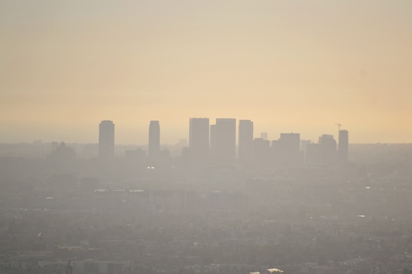 A city skyline is enveloped in dense smog during sunset, with tall buildings appearing as dark silhouettes against the hazy sky. The atmospheric conditions create a washed-out look that softens the visibility of structures in the background.