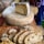 A large wheel of cheese with a wedge cut out sits on a wooden board. Several slices of rustic bread are placed on a cutting board nearby, with a knife resting beside them. The scene suggests a casual, artisanal food setting.