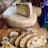 A large wheel of cheese with a wedge cut out sits on a wooden board. Several slices of rustic bread are placed on a cutting board nearby, with a knife resting beside them. The scene suggests a casual, artisanal food setting.
