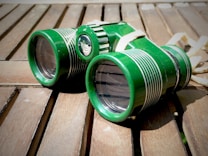 A pair of green binoculars with white detailing lies on a wooden surface, casting a shadow. The wood appears weathered and sunlight is casting bright highlights on the binoculars.