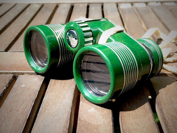 A pair of green binoculars with white detailing lies on a wooden surface, casting a shadow. The wood appears weathered and sunlight is casting bright highlights on the binoculars.