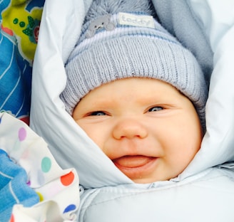 A baby is cozily wrapped in a light blue blanket, wearing a gray knitted hat with a small design and a label that reads 'teddy'. The baby has a joyful expression with a wide smile and bright eyes. In the background, a colorful patterned fabric is partially visible, adding a playful contrast.