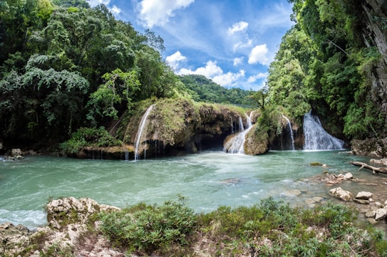 A lush, green forest surrounds cascading waterfalls that flow into a tranquil pool of blue-green water. The sky above is a bright blue, dotted with fluffy white clouds. Rocks and foliage frame the foreground, adding to the natural beauty of the scene.