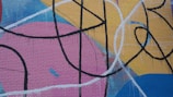 Bold graffiti artwork with abstract shapes and lines, painted on a brick wall. The design features sweeping black curves and intersecting lines over large areas of color, including pink, yellow, blue, and red.