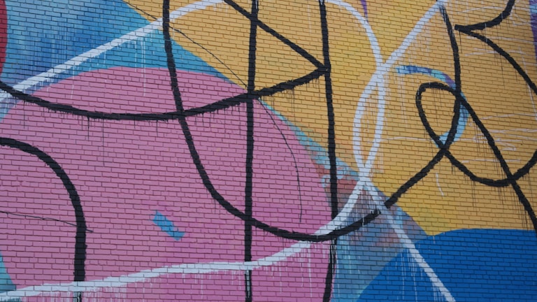 Bold graffiti artwork with abstract shapes and lines, painted on a brick wall. The design features sweeping black curves and intersecting lines over large areas of color, including pink, yellow, blue, and red.