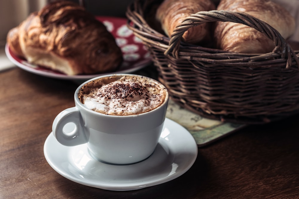 Capuccino-Kaffee auf weißer Keramiktasse mit Untertasse daneben Gebäckbrot im Korb und rotem Teller