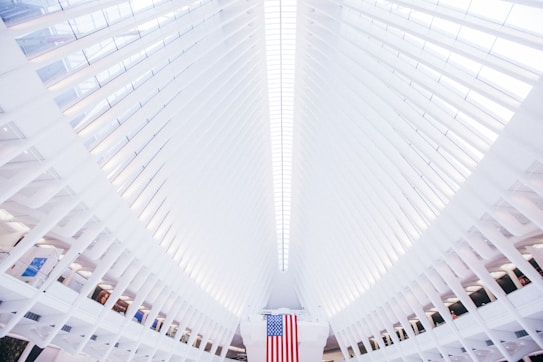 A modern architectural structure with a high ceiling and symmetrical, ribbed design. The interior features a large American flag hanging prominently in the center, with bright natural light filtering through the ceiling slats.