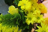 A bouquet of bright yellow flowers mixed with lush green foliage. Prominently featured are chrysanthemums and a single rose, their vibrant petals contrasting against the greenery. The arrangement appears fresh and lively.