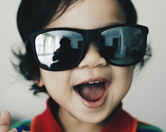 A young child with dark, curly hair is wearing oversized, black sunglasses. The child is smiling widely, displaying an expression of joy and excitement. The background is softly blurred, drawing focus to the child's facial features and accessories.