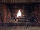 A traditional brick fireplace with a lit fire in the center. Logs arranged on a metal grate are burning, casting a warm glow. Fireplace tools are visible to the side.