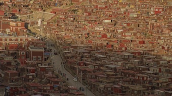 A densely packed hillside community with numerous red and brown buildings, featuring traditional architectural style. A narrow road weaves through the settlement, with a few people walking along it.