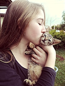A young girl with long hair is affectionately kissing a small brown tabby kitten. The kitten appears calm and is being gently held in the girl's arms. The background features outdoor greenery and part of a building.
