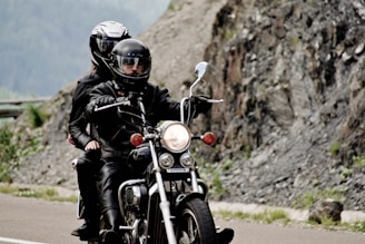 A motorcycle is being ridden by two people wearing black leather jackets and helmets. The road is winding next to a rocky landscape, with rugged mountains in the background suggesting a scenic ride.