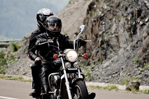 A motorcycle is being ridden by two people wearing black leather jackets and helmets. The road is winding next to a rocky landscape, with rugged mountains in the background suggesting a scenic ride.