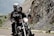 A motorcycle is being ridden by two people wearing black leather jackets and helmets. The road is winding next to a rocky landscape, with rugged mountains in the background suggesting a scenic ride.