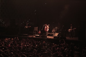 A musical band performs on stage in front of a large audience. The lead singer plays an acoustic guitar while other band members are visible with instruments like electric guitars and drums. The stage is dimly lit with a spotlight on the performers, and the crowd is lively, with hands raised and a collection of heads visible from behind.