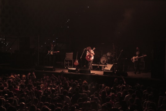 A musical band performs on stage in front of a large audience. The lead singer plays an acoustic guitar while other band members are visible with instruments like electric guitars and drums. The stage is dimly lit with a spotlight on the performers, and the crowd is lively, with hands raised and a collection of heads visible from behind.