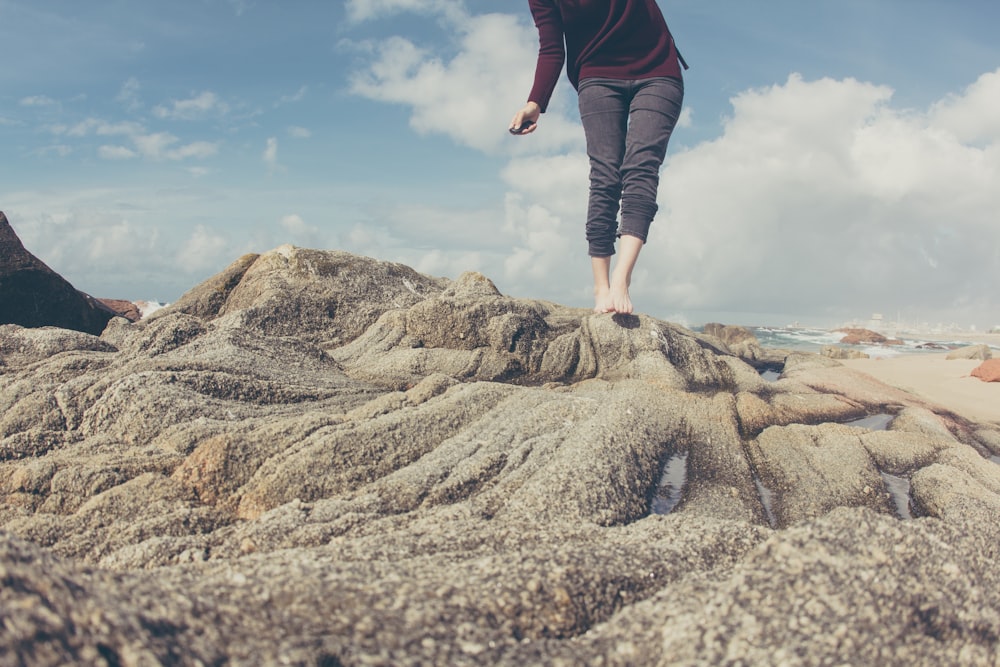 Persona che indossa pantaloni grigi in piedi sulla formazione rocciosa grigia sotto il cielo blu e bianco durante il giorno