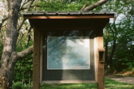 A wooden information board stands amidst a lush green setting, displaying a detailed map. The board is partially shaded by the surrounding trees, casting intricate shadows on its surface. The environment suggests a tranquil, natural park or forest trail.
