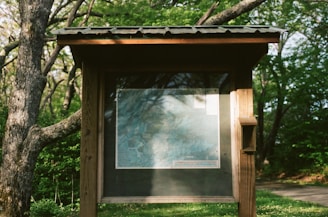 A wooden information board stands amidst a lush green setting, displaying a detailed map. The board is partially shaded by the surrounding trees, casting intricate shadows on its surface. The environment suggests a tranquil, natural park or forest trail.