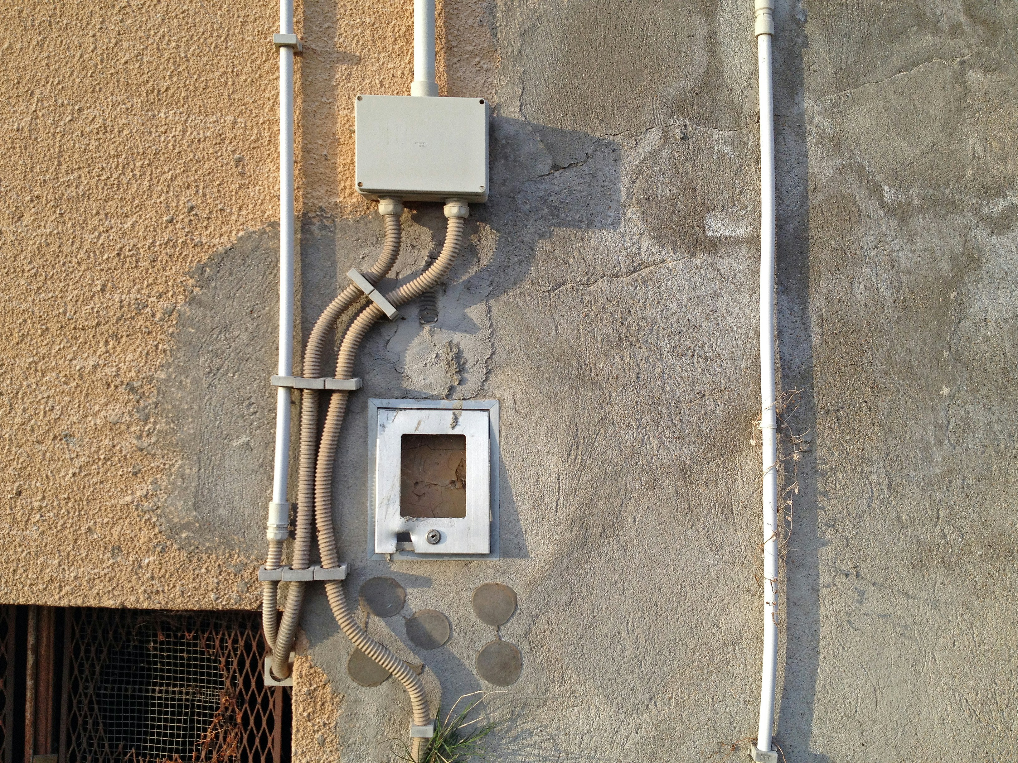 An exterior wall section showcasing various electrical conduits and a junction box attached to a textured surface. The wall has patches of paint in different shades and the sunlight casts shadows accentuating the details of the wall and conduits.
