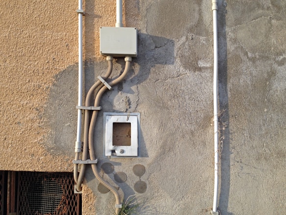 An exterior wall section showcasing various electrical conduits and a junction box attached to a textured surface. The wall has patches of paint in different shades and the sunlight casts shadows accentuating the details of the wall and conduits.