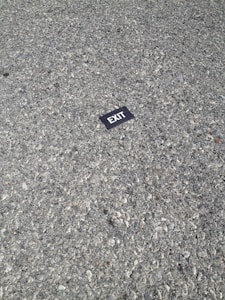 A small black rectangular sign with white text reading 'EXIT' lies on a rough, gray, pebbled asphalt surface.