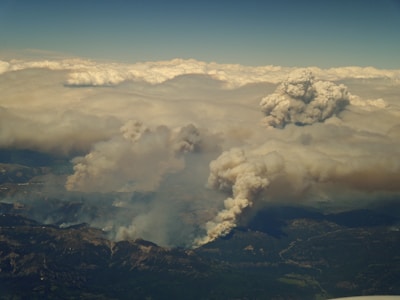 Clouds of thick smoke rise from multiple locations in a mountainous region, indicative of wildfire activity. The view suggests an aerial perspective with extensive fire coverage obscured by layers of smoke and ash. The horizon is densely packed with clouds, merging with the smoke.