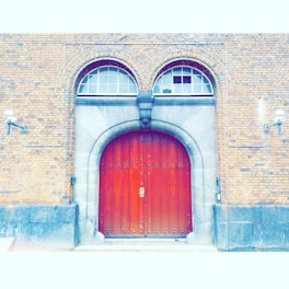 A large, wooden arched door with intricate metal hinges and a central handle is set within a stone archway. The surrounding brick wall is accentuated by two lantern-style lights and features two arched windows above the door. Electrical conduits run across the brick façade.