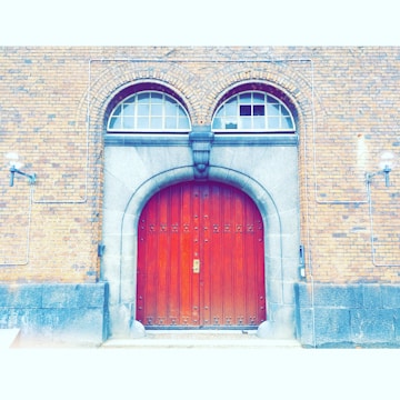 A large, wooden arched door with intricate metal hinges and a central handle is set within a stone archway. The surrounding brick wall is accentuated by two lantern-style lights and features two arched windows above the door. Electrical conduits run across the brick façade.
