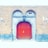 A large, wooden arched door with intricate metal hinges and a central handle is set within a stone archway. The surrounding brick wall is accentuated by two lantern-style lights and features two arched windows above the door. Electrical conduits run across the brick façade.
