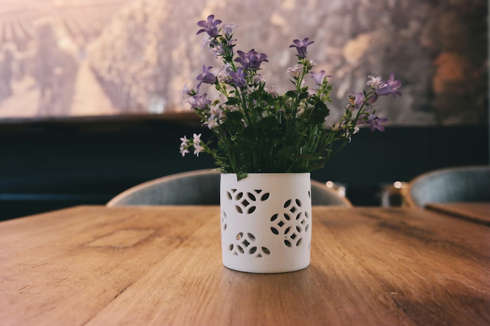 flores de pétalos púrpuras en jarrón de flores blancas ubicado en una mesa de madera marrón