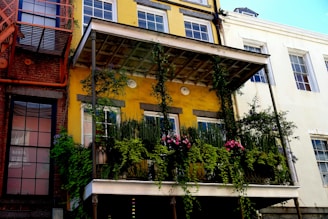 The image features a building facade with a prominent, lush balcony garden covered in green foliage and pink flowers. The building is painted in a vibrant yellow color, contrasting with the adjoining brick wall on the left and a white wall on the right. The balcony is supported by two slender metallic pillars and is adorned with a variety of plants that cascade downwards.